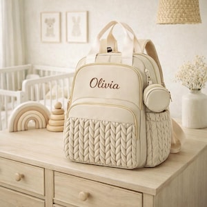 Khaki personalized diaper backpack with name Olivia in brown embroidery on beige nursery dresser, stylish baby diaper bag for parents