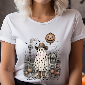 May include: A white short-sleeved t-shirt featuring a graphic print of a ghost wearing a cowboy hat and boots, holding a pumpkin balloon. The scene includes a windmill, a house, bats, and the text "boo y'all" in a black cursive script.