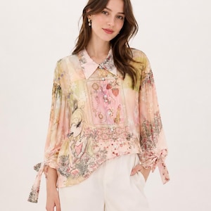 Peut inclure: Un chemisier transparent à manches longues avec un imprimé floral et de dessins animés dans des tons de rose, jaune et vert. Le chemisier présente un col, des poignets noués et est associé à un pantalon blanc.