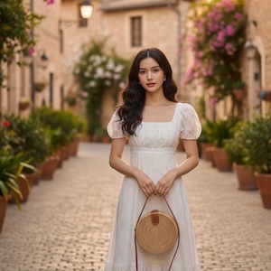 May include: A white midi dress with a square neckline and short, puffed sleeves. The dress features a detailed lace hem and is paired with a round, woven handbag with a leather strap. The scene is set on a cobblestone street.