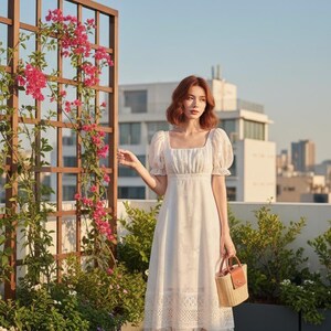 May include: A white midi dress with a square neckline and short, puffed sleeves. The dress features a floral pattern and a lace hem. A small woven handbag with a brown leather handle is held. The background includes a wooden trellis with pink flowers.
