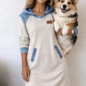 Puede incluir: Un vestido con capucha color crema con detalles de mezclilla, incluyendo la capucha, los puños y los detalles de los bolsillos. El vestido presenta un pequeño parche marrón y lo lleva una persona que sostiene un perro corgi.