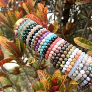 May include: A collection of beaded bracelets in various colors, including green, black, purple, and pink. The bracelets are displayed against a backdrop of colorful foliage, showcasing the diverse hues of the gemstones.