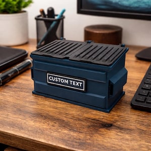 May include: A navy blue miniature dumpster with a black lid and the text "CUSTOM TEXT" on a white rectangle. The miniature dumpster is sitting on a wooden desk.