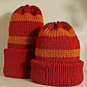May include: Two knitted beanies featuring a ribbed texture and a cuffed design. The hats are patterned with horizontal stripes in alternating shades of deep red and bright orange, constructed with a thick yarn in a consistent knit stitch pattern.