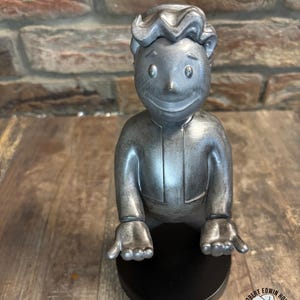 May include: A silver-colored figurine of a smiling cartoon character with a black base. The figurine has a classic hairstyle and is wearing a suit. The base has the text "MR ROBERT EDWIN HOUSE'S WASTELAND ODDITIES" and a cartoon image.
