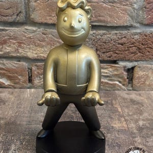 Könnte beinhalten: Goldfarbene Figur einer Zeichentrickfigur mit lächelndem Gesicht, die auf einem schwarzen, kreisförmigen Sockel steht. Die Figur hat ein Retro-Design und ist mit offenen Händen posiert. Der Sockel trägt den Text "Mr. Robert Edwin House's Wasteland Oddities".