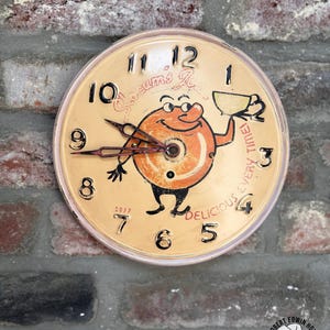 May include: A vintage wall clock with a cream-colored face featuring a cartoon character holding a teacup. The clock has black numerals, hands, and the text "Delicious Every Time!" The clock is mounted on a brick wall.