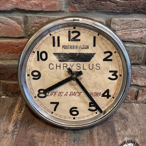May include: A round wall clock with a metallic silver frame and a cream-colored face. The clock features black numerals, black hands, and the text "POWERED BY FUSION," "CHRYSLUS," and "LIFE IS A RACE... WIN!" printed in black on the dial.