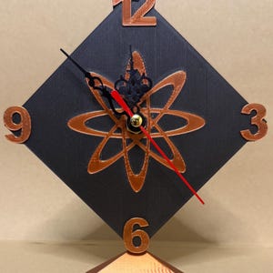 May include: A diamond-shaped black clock featuring copper-colored numbers 12, 3, 6, and 9. The center displays a copper-colored atomic symbol design. The clock has ornate black hour and minute hands, a thin red second hand, and a copper base.