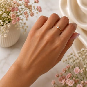 Puede incluir: Un anillo fino de oro con una pequeña perla blanca única centrada en la banda. El anillo se lleva en el dedo corazón y presenta un diseño minimalista con un acabado metálico pulido y una perla blanca redonda, lisa y brillante.