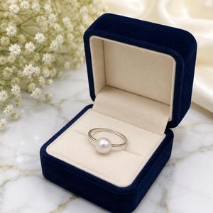 Könnte beinhalten: Ein silberner Ring mit einer einzelnen runden weißen Perle in einer marineblauen Samtschmuckschatulle mit cremefarbenem Innenfutter. Der glatte, metallische Ringband hält die Perle sicher in der Mitte des geöffneten Schmuckkästchens.