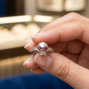 May include: A silver skull ring with red gemstone eyes. The ring is held up, showcasing the detailed skull design and the decorative band. The ring is a gothic-inspired piece of jewelry.