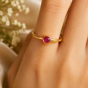 Könnte beinhalten: Ein dünner Goldring mit einem einzelnen, runden, facettierten, tiefrosa Rubin, der in einer glatten Zargenfassung sitzt. Die Ringschiene ist poliert und schmal und zeigt ein schlichtes Design, das die leuchtende Farbe des Steins betont.