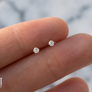 May include: A pair of small diamond stud earrings with a gold setting. The earrings are shown on a finger. Each earring features a single, sparkling diamond. The design is classic and elegant, suitable for everyday wear or special occasions.