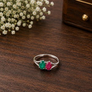 Puede incluir: Un anillo de tono plateado con dos piedras preciosas de corte ovalado, una de color verde vibrante y otra de color rojo intenso, engastadas una al lado de la otra en una banda de vástago dividido. El anillo se muestra sobre madera oscura.