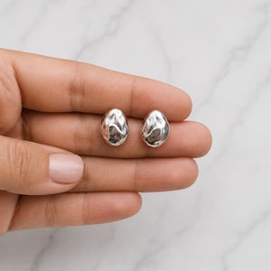 Può includere: Un paio di piccoli orecchini ovali in argento con una superficie lucida, irregolare e martellata. Gli orecchini presentano una finitura metallica riflettente e una forma organica arrotondata che cattura la luce sulla loro superficie irregolare e ammaccata.