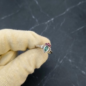 Op de afbeelding: Een zilveren ring met een peervormige, smaragdgroene edelsteen en drie kleine, ronde, rode edelstenen. De ring heeft een gedetailleerde, kralenband en een decoratieve zetting. De ring wordt door een gehandschoende hand tegen een donkere achtergrond gehouden.