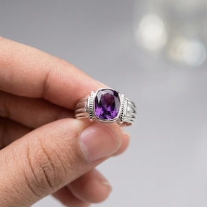 Pode incluir: Um anel de prata com uma grande pedra preciosa roxa de forma oval. O anel tem uma banda detalhada com linhas horizontais e pequenos elementos decorativos nas laterais da pedra preciosa. O anel é segurado por uma mão.