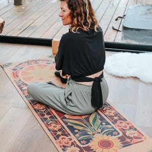 May include: A yoga mat with a vibrant floral and geometric pattern. The mat displays a large sunflower at its centre, encircled by flowers and shapes in red, orange, blue, and green. The word "Amsom&eacute;" is printed on the mat.
