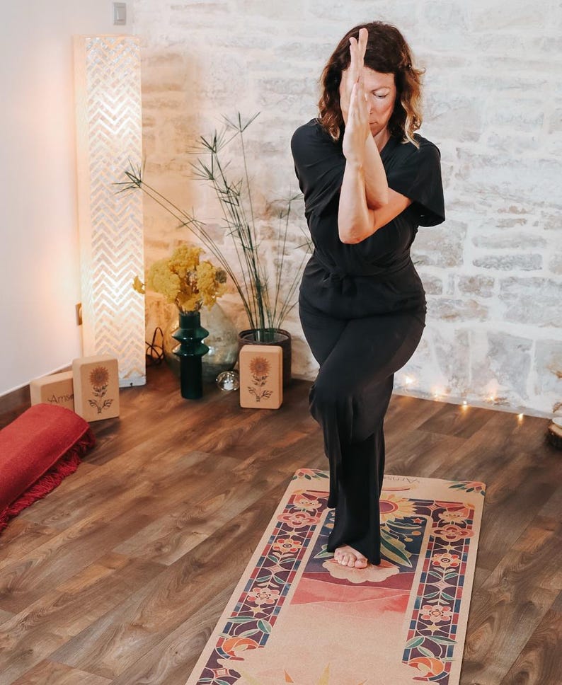 May include: A person in black clothing performs a yoga pose on a patterned yoga mat. The mat has a floral design with a sunflower in the middle. The background includes a stone wall, plants, and wooden blocks.