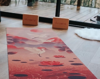 May include: A yoga mat with a tranquil design featuring white birds, red lotus flowers, and a water scene in shades of red and pink. Two wooden yoga blocks with the word "Amseme" are at the top. The mat is on a wooden floor.