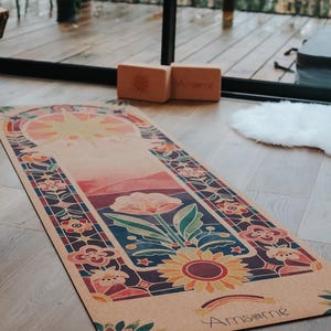 May include: A rectangular yoga mat with a colourful floral design, featuring a large sunflower and a rainbow. The mat has a light brown background with a stained-glass style border. Two wooden yoga blocks are visible.