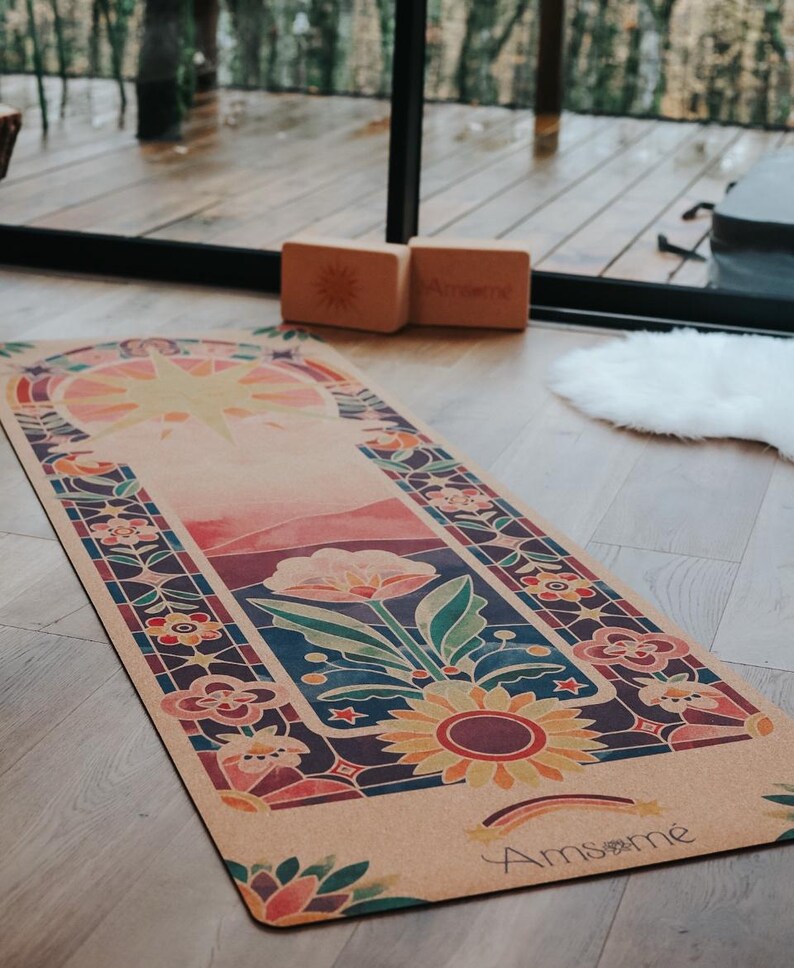 May include: A rectangular yoga mat with a colourful floral design, featuring a large sunflower and a rainbow. The mat has a light brown background with a stained-glass style border. Two wooden yoga blocks are visible.