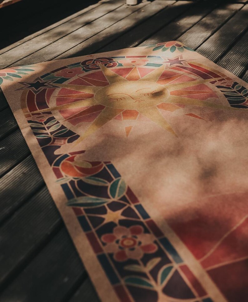 May include: A yoga mat featuring a sun design in yellow, orange, red, and blue. The mat has a stained glass style border with floral and star motifs. The mat is laid out on a wooden deck.