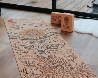 May include: A tan yoga mat with a nature-themed illustration featuring a large sunflower, mushrooms, and leaves. The mat has a mountain and cloud design and the word "Amsome" printed on the bottom. Two wooden yoga blocks are also visible.