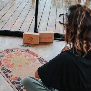 May include: A yoga scene with a colourful yoga mat featuring a sun design, two wooden blocks with the text "Amsome", and a person in a black top and green trousers. The mat has a sun design with a rainbow of colours.