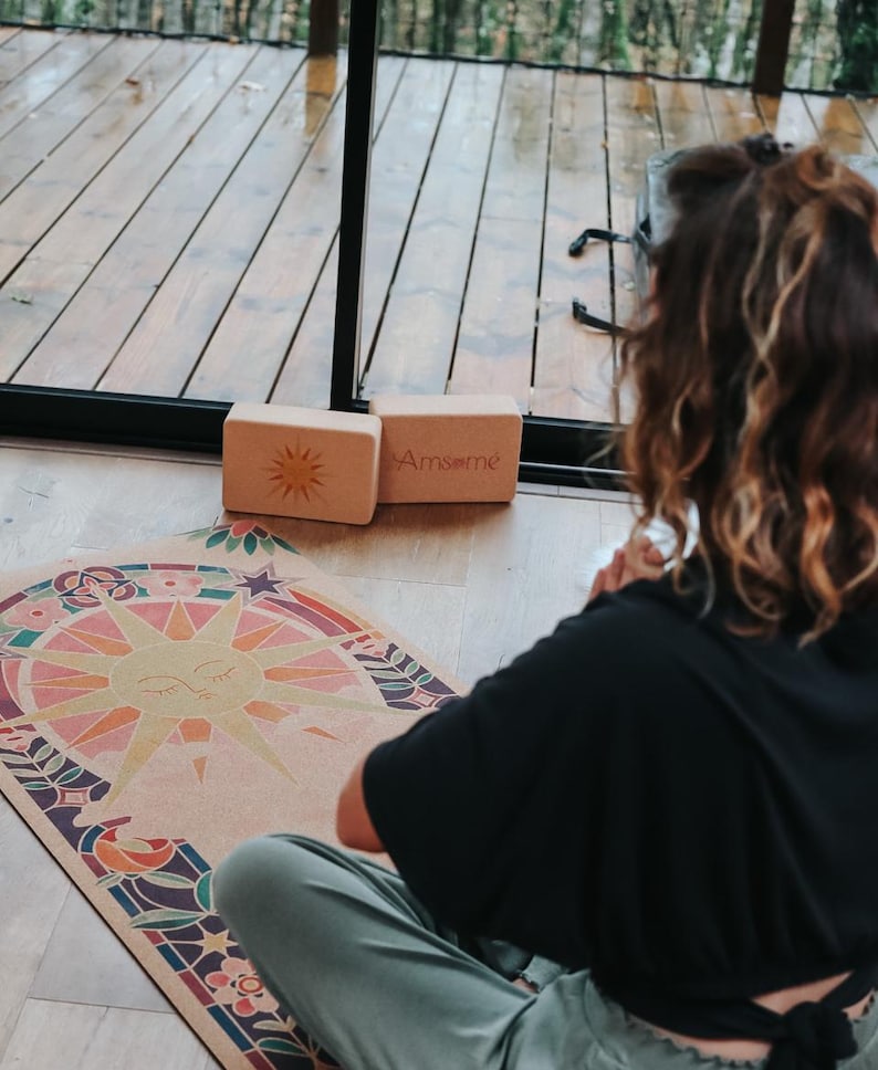 May include: A yoga scene with a colourful yoga mat featuring a sun design, two wooden blocks with the text "Amsome", and a person in a black top and green trousers. The mat has a sun design with a rainbow of colours.