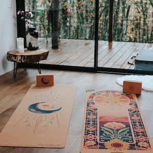 May include: Two yoga mats with celestial designs and wooden yoga blocks are displayed on a wooden floor. One mat features a crescent moon and the other a sunburst design. The mats are tan with colourful illustrations.
