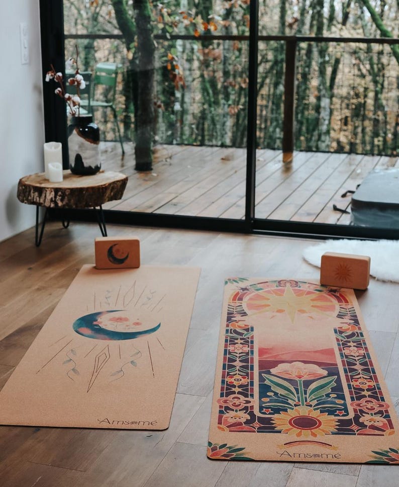 May include: Two yoga mats with celestial designs and wooden yoga blocks are displayed on a wooden floor. One mat features a crescent moon and the other a sunburst design. The mats are tan with colourful illustrations.