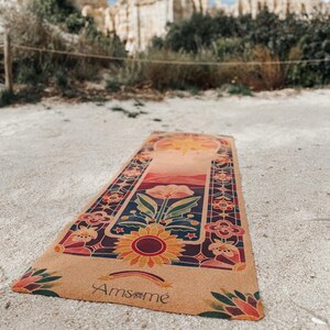 May include: A rectangular yoga mat with a floral design featuring a sunflower, flowers, and a rainbow. The mat has a tan background with a colourful border and the word "Amsm&eacute;" printed on it. The mat is outdoors on a sandy surface.
