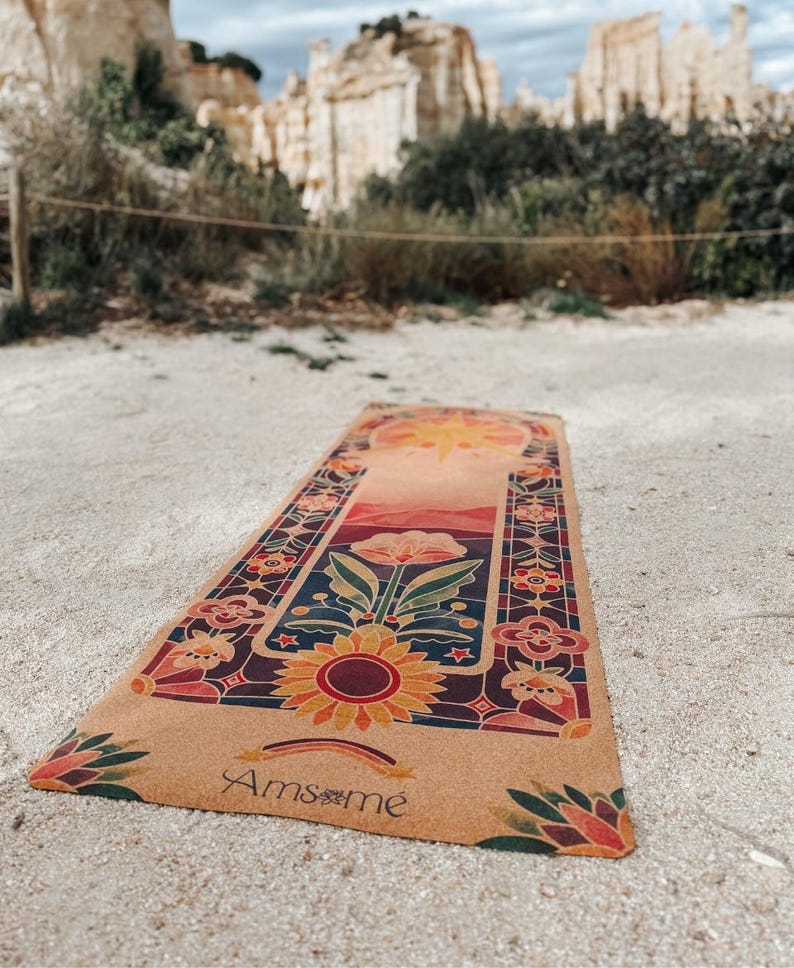 May include: A rectangular yoga mat with a floral design featuring a sunflower, flowers, and a rainbow. The mat has a tan background with a colourful border and the word "Amsm&eacute;" printed on it. The mat is outdoors on a sandy surface.