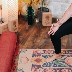 May include: A yoga mat with a colorful floral design featuring a large sunflower and the word "Amsam&eacute;". A rolled-up red yoga mat with fringe is next to the mat. Yoga blocks with sunflower designs are also visible.
