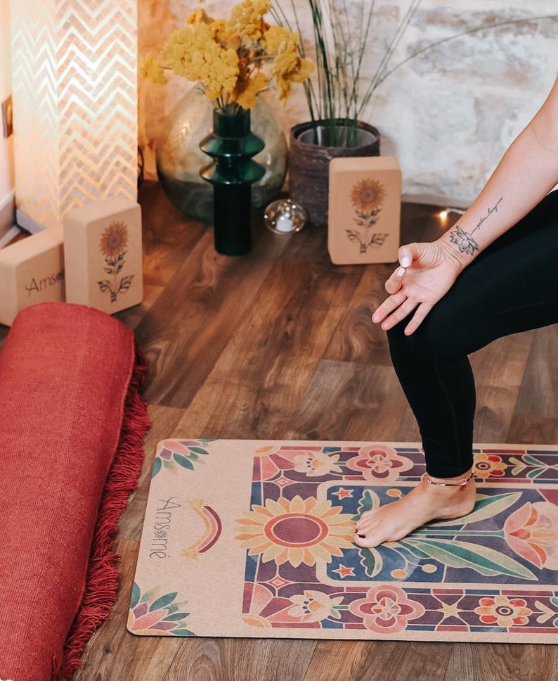 May include: A yoga mat with a colorful floral design featuring a large sunflower and the word "Amsam&eacute;". A rolled-up red yoga mat with fringe is next to the mat. Yoga blocks with sunflower designs are also visible.