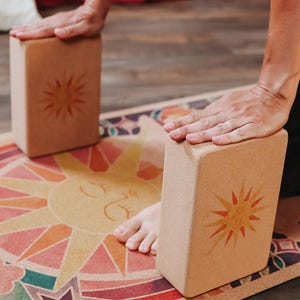 May include: Two rectangular yoga blocks with a sunburst design are positioned on a colourful yoga mat. The blocks are light brown and the mat features a sun graphic and geometric patterns in various colours. The blocks are being used for support.