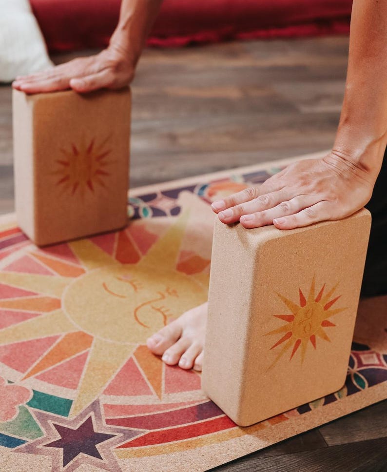 May include: Two rectangular yoga blocks with a sunburst design are positioned on a colourful yoga mat. The blocks are light brown and the mat features a sun graphic and geometric patterns in various colours. The blocks are being used for support.