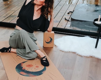 May include: A yoga mat featuring a crescent moon and floral design, alongside a matching yoga block. The mat is tan with a blue, pink, and orange moon design. The yoga block is light brown with a crescent moon design.