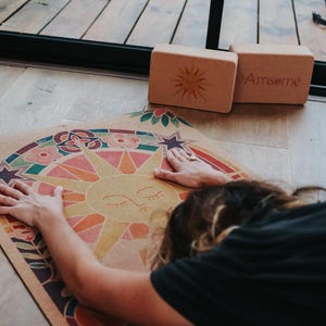 May include: A yoga mat featuring a colourful sun design and a person stretching on it. Two rectangular yoga blocks are nearby. One block has a sun design, and the other has the text "Amsome". The mat and blocks are on a wooden floor.
