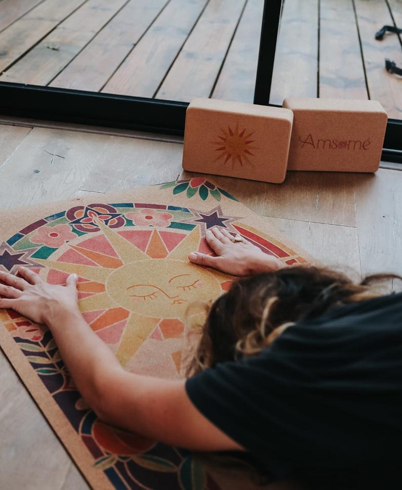 May include: A yoga mat featuring a colourful sun design and a person stretching on it. Two rectangular yoga blocks are nearby. One block has a sun design, and the other has the text "Amsome". The mat and blocks are on a wooden floor.