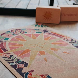 May include: A yoga mat with a colourful sun design, surrounded by floral and geometric patterns. Two wooden yoga blocks are in the background. The mat features a beige background with a sun graphic in shades of pink, orange, and yellow.