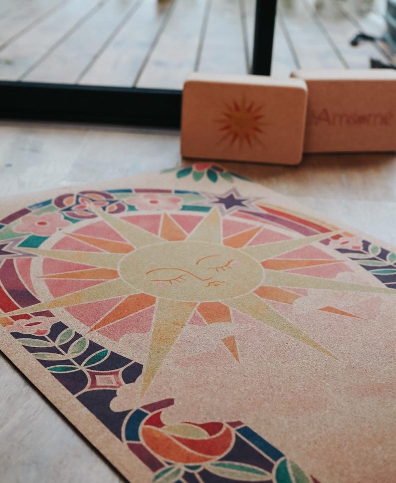 May include: A yoga mat with a colourful sun design, surrounded by floral and geometric patterns. Two wooden yoga blocks are in the background. The mat features a beige background with a sun graphic in shades of pink, orange, and yellow.