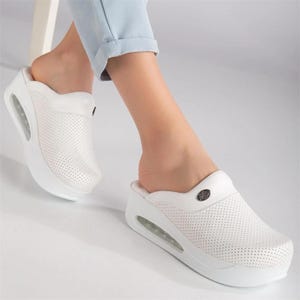 May include: White perforated clogs with a thick sole and air cushion. The clogs have a rounded toe and a small decorative emblem on the top. The shoes are shown on a person's feet.