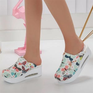 Könnte beinhalten: Weiße Clogs mit einem Schmetterlingsmuster in Rosa, Blau und Grün. Die Clogs haben eine dicke weiße Sohle mit Luftpolster. Die Schuhe sind hinten offen und wirken bequem.