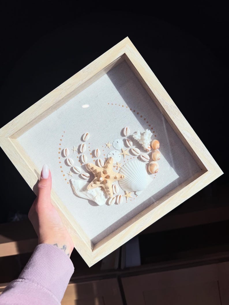 May include: A framed seashell art piece featuring a starfish, various seashells, and coral arranged on a light-colored fabric. The arrangement is enclosed in a natural wood frame, creating a coastal theme.