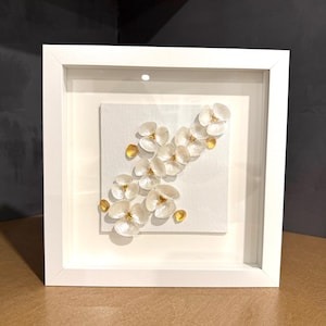Könnte beinhalten: Ein quadratischer weißer Rahmen mit einem weißen Stoffkunstwerk. Das Kunstwerk zeigt eine Ansammlung kleiner, irisierender weißer Muschelblüten, die diagonal angeordnet sind und in der Mitte jeder Blüte mit winzigen, gelben Perlen verziert sind.