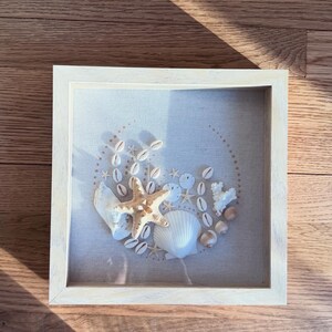 May include: A framed beach-themed artwork featuring a collection of seashells, starfish, and coral arranged on a light-colored fabric. The arrangement is enclosed in a square, light-wood frame, creating a coastal aesthetic. The artwork is displayed on a wooden surface.